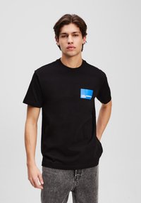 Melnas kokvilnas t-krekls ar apaļu kaklu, īsām piedurknēm, ar zilu taisnstūrveida kabatu ar baltu tekstu. Brīva piegriezuma stils.