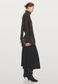 Green knit sleeveless top over a brown long-sleeve shirt, black pleated skirt, and knee-high black leather boots. Holding a woven brown bag.