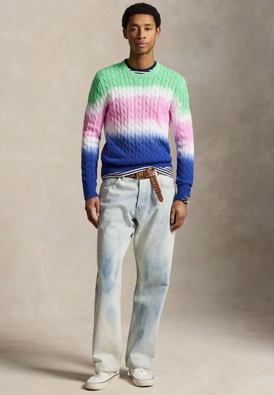 Homme debout portant un pull torsadé rayé vert, rose, blanc et bleu, un jean clair, une ceinture tressée marron et des chaussures blanches.