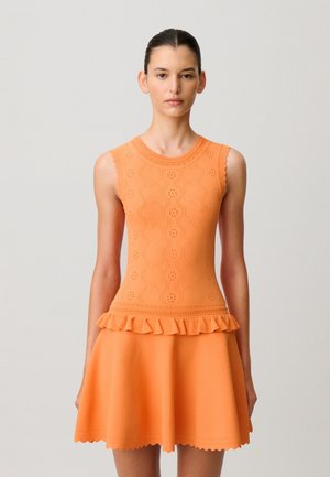 Mujer con cabello oscuro recogido en un moño, vistiendo una blusa sin mangas de punto naranja con patrones florales y borde con volantes, combinada con una falda naranja a juego.