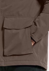 Bruine waterdichte jas met een grote klepzak, textuur op het oppervlak en klittenbandsluitingen bij de mouwen voor een stevige pasvorm.