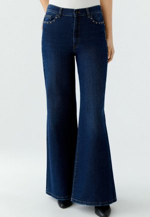 Donkerblauwe flared jeans met een hoge taille, voorzien van voorzakken en decoratieve studs langs de rand van de zakken. Gemaakt van denim stof.