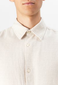 Light beige button-up shirt with a textured, ribbed fabric. Features a classic collar and pearl-like buttons.