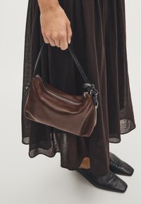 Brown leather handbag with a slim shape, featuring a zipper closure and a black strap. Set against a dark outfit, showcasing its texture.