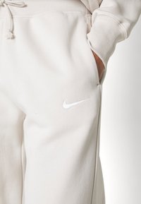 Gai bēšā krāsā izgatavotas jogger stila bikses no mīksta auduma, ar baltu Nike logo, ribētām piedurknēm un aukliņu jostu. Malās ir kabatas.