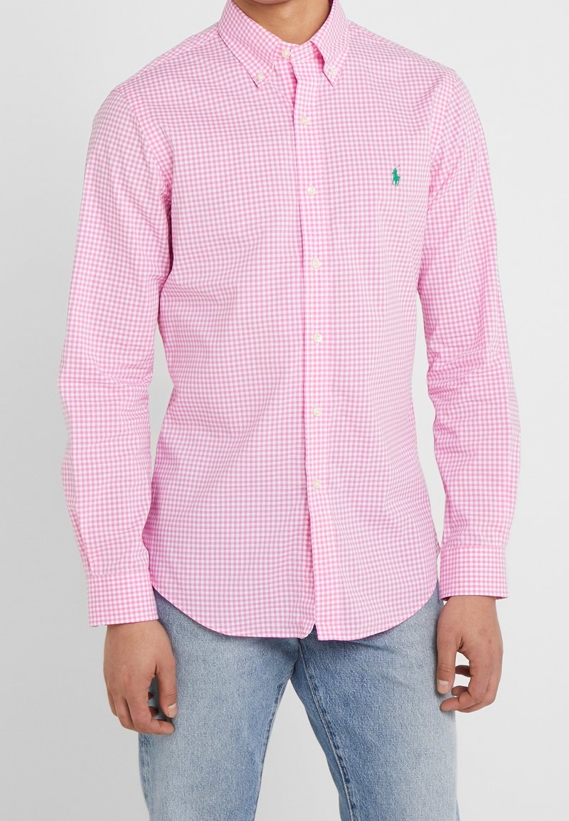 Růžová kostkovaná košile na knoflíky s dlouhými rukávy, klasickým límečkem a malým zeleným logem na levé straně hrudníku. Materiál je hladký a lehký.