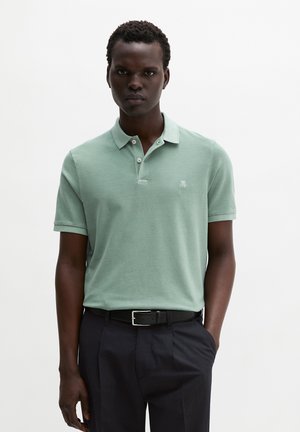 Homme portant un polo à manches courtes vert clair rentré dans un pantalon noir avec une ceinture noire, debout devant un fond uni.