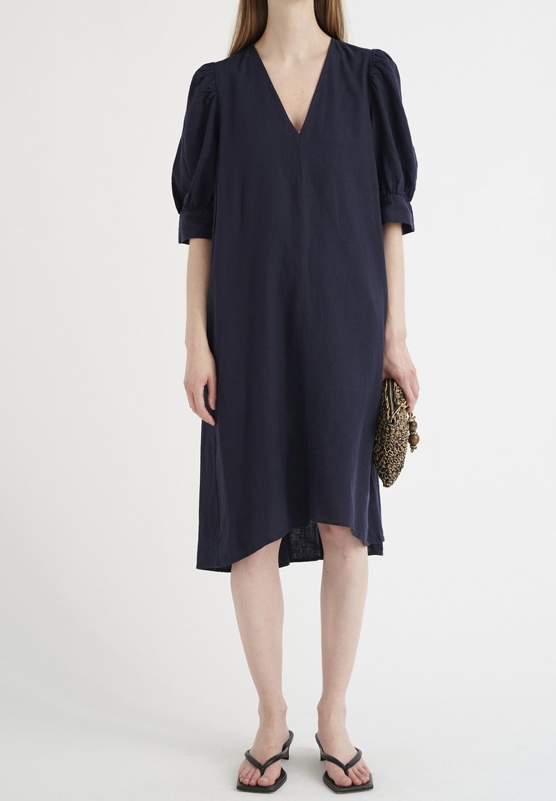 Granatowa lniana sukienka z dekoltem w kształcie litery V, z bufiastymi rękawami i asymetrycznym dołem; zestawiona z klapkami i plecioną torebką.
