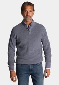 Grijze gebreide pullover met een hoge kraag, voorzien van twee groene knopen en rode en witte accenten op de manchetten. Draagt het met een blauwe spijkerbroek.
