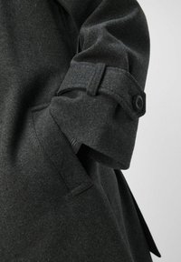 Manche de manteau en laine gris foncé présentant un poignet boutonné, un design ajusté et une texture de tissu structuré avec des détails de couture visibles.