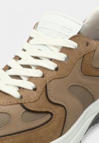 Καφέ σουέντ και δίχτυνο αθλητικό παπούτσι με λευκά κορδόνια, διαθέτοντας υφασμάτινο σχέδιο και μαξιλαρωμένη γλώσσα, διακοσμημένο με λογότυπο σε τόνους.