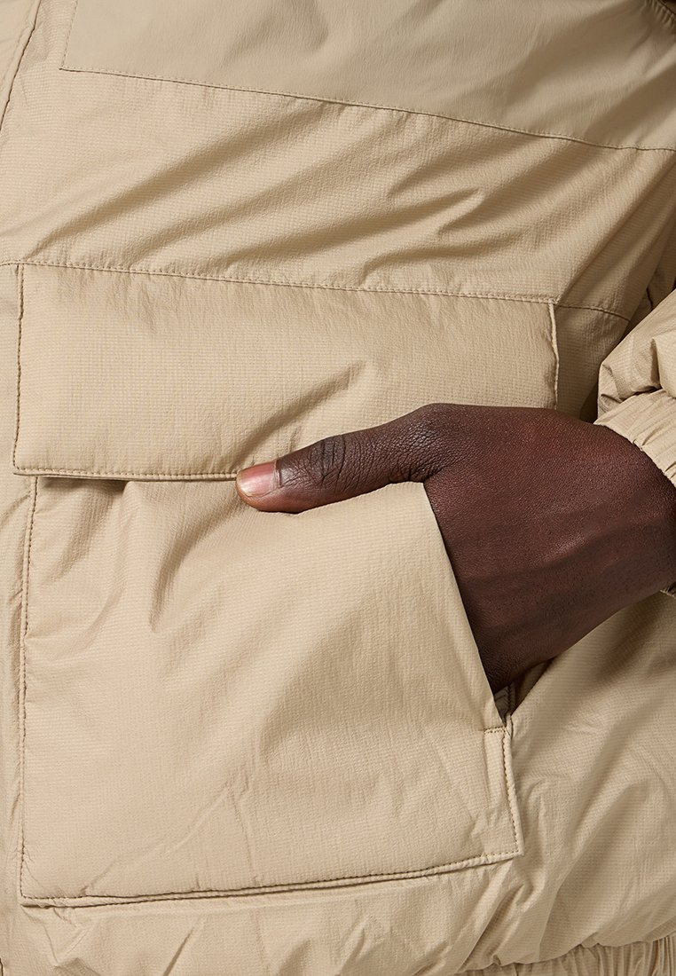 Chaqueta acolchada beige con un tejido texturizado, que presenta un gran bolsillo de parche en la parte frontal y una mano parcialmente insertada en el bolsillo.