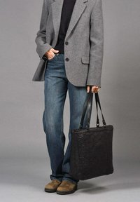 Zwarte gestructureerde tote bag met dubbele handgrepen, gedragen bij een grijze blazer en blauwe jeans, staande op een neutrale achtergrond.