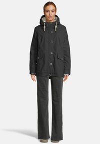 Vrouw in een zwarte regenjas met capuchon en knoopsluiting, met voorflappen en wijde, donkergrijze broek, staand op een witte achtergrond.