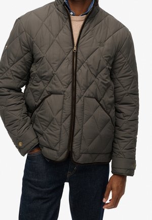 Hombre con chaqueta acolchada de color verde oliva oscuro cerrada con cremallera sobre un suéter beige y una camisa azul, combinada con pantalones vaqueros oscuros.