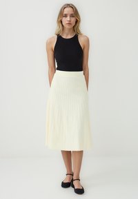 Top sans manches noir associé à une jupe plissée légère de couleur jaune pâle, arrivant au genou. Texture lisse avec de petites lignes verticales. Chaussures plates noires.