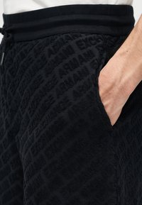 Gros plan sur une main dans la poche d'un short noir texturé avec le texte "Armani Exchange" en relief répété et une ceinture côtelée.