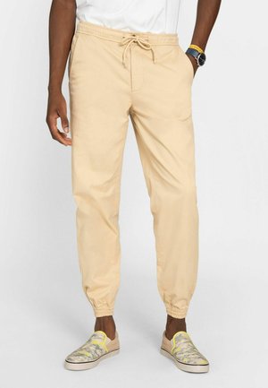 Mand iført beige joggerbukser med snøre og camouflage slip-on sko, med den ene hånd i lommen og en hvid skjorte stoppet ned.