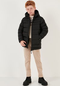 Zwarte pufferjack met een capuchon, voorzien van een horizontaal gestikt patroon, ritszakken en een rechte snit, gecombineerd met beige broek en zwarte laarzen.