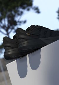Par sorte løbesko med Cordura stofetiket hviler på en hvid overflade udendørs mod en sløret træbaggrund.