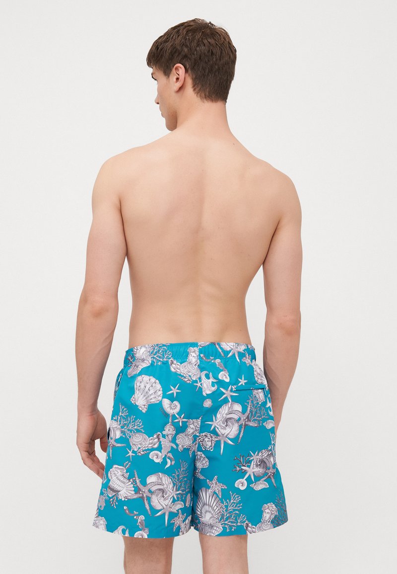 Blå svøm shorts med et koral- og muslingmønster, med elastisk talje og snor, vist fra bagsiden.