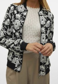 Kurtka bombers z kwiatowym wzorem na czarnym tle, z białymi wzorami piwonii, ściągaczami przy mankietach i zapięciem na zamek. Noszona na teksturowanej białej bluzce.