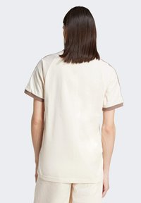 Φωτεινό μπεζ μπλουζάκι με στρογγυλή λαιμόκοψη, κοντά ραγκλάν μανίκια με καφέ λεπτομέρειες, και μαλακή βαμβακερή υφή, εμφανίζεται από πίσω.