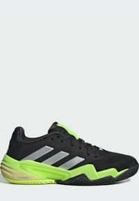 Zapatilla deportiva negra con acentos verdes, parte superior de malla texturizada, tres franjas plateadas y una suela gruesa con un patrón de agarre único.