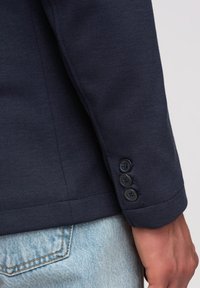 Ärmel eines Marineblazers mit drei Knöpfen, der über der Tasche einer hellblauen Jeans liegt, und eine Hand daneben.