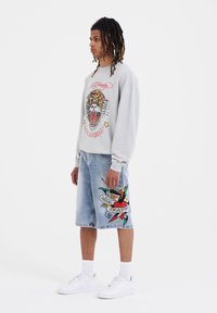 Felpa grigia chiara con grafica di tigre e dettagli rossi, abbinata a pantaloni corti in denim blu con un design colorato in stile tatuaggio. Sneakers bianche.
