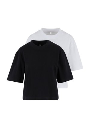 Femme aux cheveux bouclés portant un t-shirt noir ample et un jean bleu clair, debout avec une main dans la poche devant un fond uni.