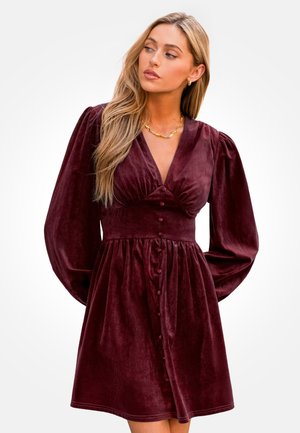 Bourgondische fluwelen jurk met V-hals, pofmouwen en een knoopsluiting aan de voorkant. Zachte textuur en een geraffineerde taille die zorgt voor een uitlopende silhouet.