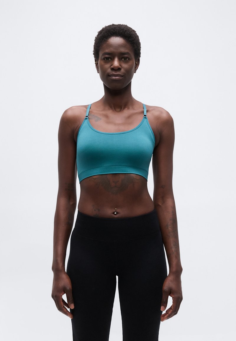 Αθλητική γυναίκα που στέκεται με το πρόσωπο προς τα εμπρός, φοράει ένα τυρκουάζ αθλητικό σουτιέν και μαύρο κολάν, με φόντο έναν απλό φωτεινό τοίχο.