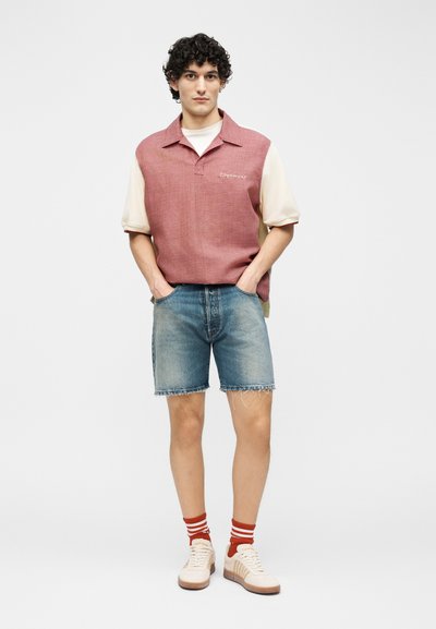 Jeune homme portant un gilet tricoté rouge sans manches sur une chemise beige, un short en denim, des chaussettes rayées rouges et des baskets beiges, les mains dans les poches.