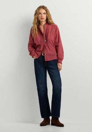 Frau mit langen lockigen Haaren, die eine rote, geknöpfte Bluse, dunkelblaue Jeans, braune Knöchelstiefel und eine Anhänger-Halskette trägt, steht vor einem schlichten Hintergrund.