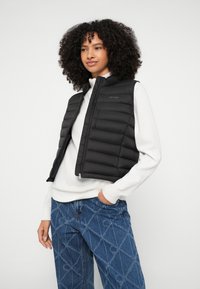 Μαύρο μονωμένο αμάνικο γιλέκο με σχεδιασμό καπιτονέ, συνδυασμένο με λευκό πουκάμισο μακριά μανίκια και τζιν με γεωμετρικό σχέδιο.