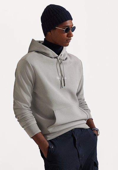 Sweatshirt cinza claro com bolso frontal, capuz com cordão e logótipo bordado. Usado com um gorro azul-marinho canelado e calças escuras.