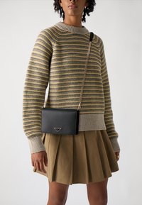 Svart lærcrossbody-veske med kjedehank, med en trekantet aksent. Modellen har på seg en stripete genser og en plisserte khaki-skjørt.