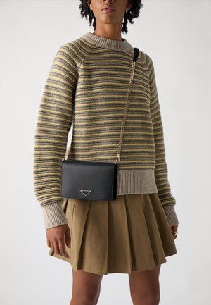 Borsa a tracolla nera in pelle con tracolla a catena, caratterizzata da un accento triangolare. Il modello indossa un maglione a righe e una gonna plissettata khaki.