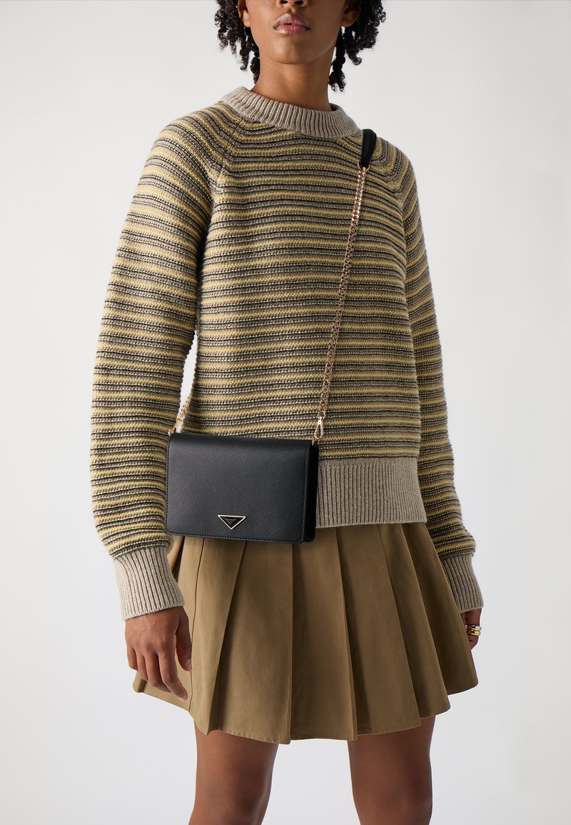 Svart lærcrossbody-veske med kjedehank, med en trekantet aksent. Modellen har på seg en stripete genser og en plisserte khaki-skjørt.