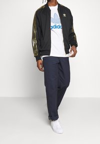 Svart zip-up jacka med guldstripade detaljer, vit t-shirt med blå logotyp, marinblå byxor och vita sneakers. Avslappnad sportig stil.