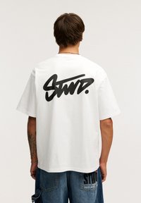 Hvid bomulds T-shirt med korte ærmer, der har en stor sort grafisk tekst "STWD." på ryggen. Enkel design og almindelig pasform.
