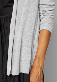 Szary dziergany kardigan w luźnym kroju, z prążkowanymi mankietami i miękką fakturą, nałożony na czarną plisowaną sukienkę.