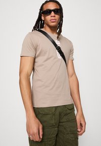 Beige bomuld t-shirt med korte ærmer, prydet med et centralt logo-patch. Kombineret med olivengrønne cargo-bukser, som er accentueret med flere lommer.