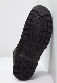 Suela de bota de goma negra con profundas estrías, un patrón texturizado y un logotipo. El diseño es resistente, con una forma redondeada y materiales duraderos.