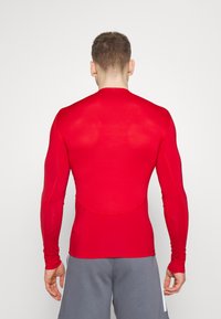 Long-sleeve athletic shirt in bright red, made of stretchy fabric. Features fitted design and contrasting seam accents.