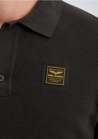 Zwarte polo met een gestructureerde textuur, voorzien van een knoopkraag en een vierkant label met gouden en zwarte tekst: "PME LEGEND TESTED AT HIGH ALTITUDE."