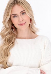 Sweater blanc en tricot avec un col large et une texture côtelée. Cheveux longs et ondulés et grandes boucles d'oreilles dans des tons de rouge et de rose.