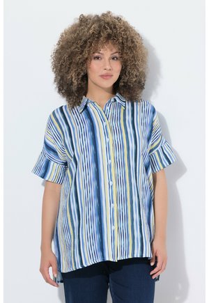 Kvinde med krøllet hår iført en løs, kortærmet skjorte med lodrette blå, gule og hvide striber, stående mod en hvid baggrund.