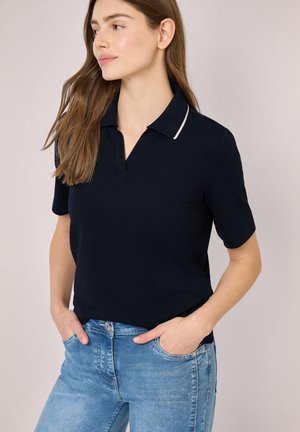Femme aux longs cheveux bruns portant un polo noir à manches courtes et un jean bleu clair, les mains dans les poches, regardant vers la gauche.